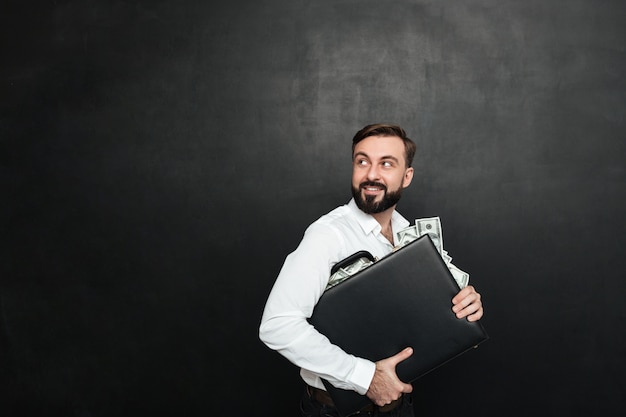 Portret van gelukkig rijk zakenman dragend aktentashoogtepunt van dollarrekeningen en terug kijkend, geïsoleerd over donkergrijs