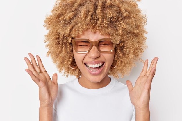Portret van gelukkige blije vrouw met krullend borstelig haar houdt handen omhoog grijns op camera reageert op positief nieuws draagt bril en casual t-shirt geïsoleerd op witte achtergrond Menselijke emoties