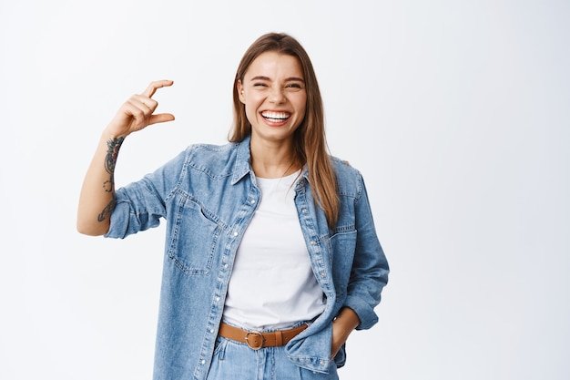 Portret van gelukkige blonde vrouw in stijlvolle kleding, lachend met gesloten ogen en witte glimlach, klein formaat met vingers tonend, klein ding vormgevend, staande over witte muur