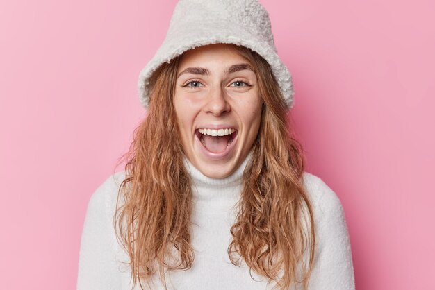 Portret van gelukkige dolblije vrouw roept luid voelt vrolijk heeft aangename uitstraling voelt vrolijk gekleed in warme witte trui en panama geïsoleerd over roze achtergrond. Mensen en emoties