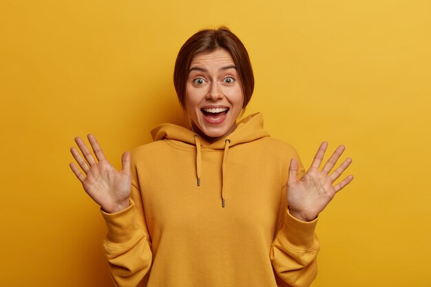 Portret van gelukkige jonge Europese vrouw glimlacht positief, werpt handpalmen op en kijkt graag, verwacht niet zo leuk cadeau te krijgen, draagt casual gele hoodie in één toon met muur