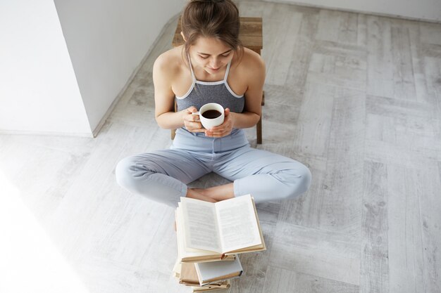 Portret van gelukkige jonge mooie vrouw het glimlachen de kop van de het boekholding van het lezingsboek van koffiezitting op vloer over witte muur.