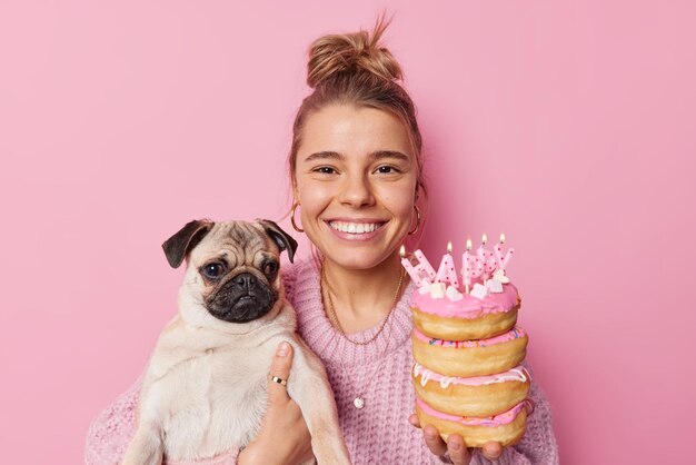 Portret van gelukkige jonge mooie vrouw poseert met pug dog viert huisdieren verjaardag houdt stapel heerlijke donuts met brandende kaarsen geïsoleerd over roze achtergrond. Speciale gelegenheid concept