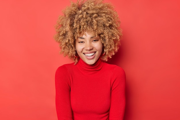 Portret van gelukkige jonge vrouw glimlacht breed draagt casual coltrui voelt blij poses blij tegen rode achtergrond. Vrolijk vrouwelijk model heeft een vrolijke uitdrukking, voelt zich optimistisch en geniet van een goede dag