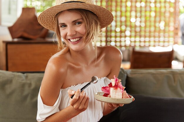 Portret van gelukkige jonge vrouw komt op zomerfeest, iets speciaals vieren, gekleed in zomerkleding, heerlijke cake eet, heeft een blijde positieve uitdrukking. Mensen, eten en rusten concept