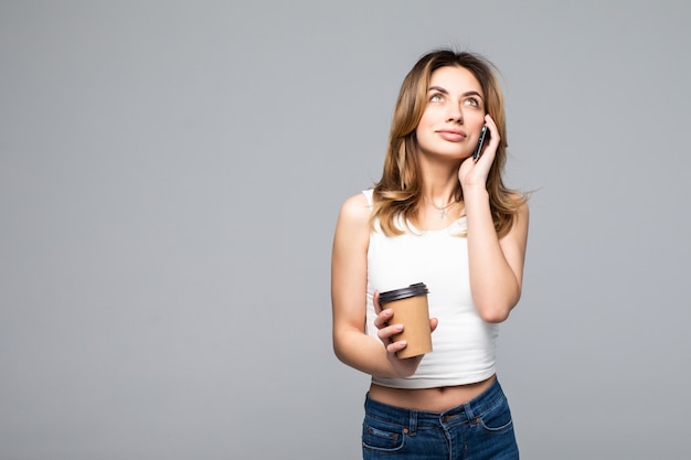 Portret van gelukkige jonge vrouw poseren praten door mobiele telefoon drinken koffie geïsoleerd over grijze muur