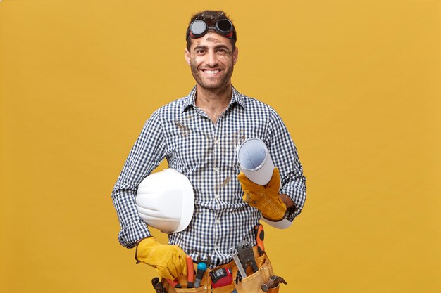 Portret van gelukkige mannelijke werknemer in vrijetijdskleding, het dragen van beschermende brillen, handschoenen en het hebben van gereedschapsriem op de taille met blauwdruk en helm met aangename glimlach verheugend op zijn succes op het werk
