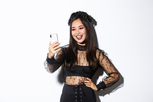 Portret van gelukkige mooie Aziatische vrouw in halloween kostuum glimlachen en kijken naar het scherm van de mobiele telefoon, met video-oproep, staande over witte muur