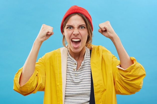 Portret van gelukkige succesvolle jonge blanke vrouwenwinnaar die rode hoed en gele regenjas draagt die zich verheugt over overwinning, succes of goed positief nieuws met gebalde vuisten, juichend, schreeuwend van vreugde
