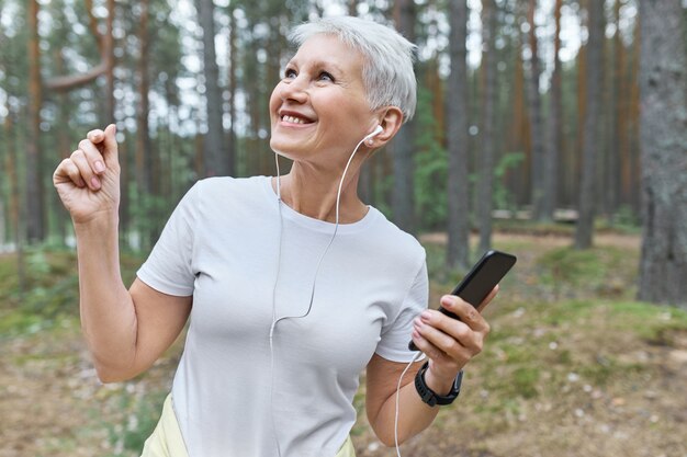 Portret van gelukkige vrolijke volwassen vrouw in wit t-shirt en oortelefoons met plezier buitenshuis