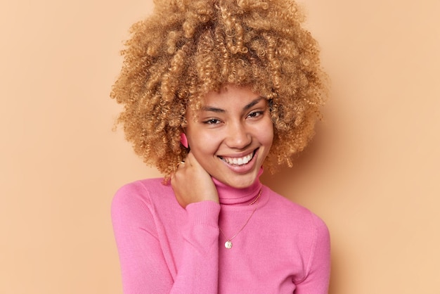 Portret van gelukkige vrolijke vrouw houdt hand op nek glimlacht graag heeft natuurlijk krullend haar gezonde huid voelt gelukkig draagt roze poloneck geïsoleerd over bruine achtergrond. Mensen en emoties concept