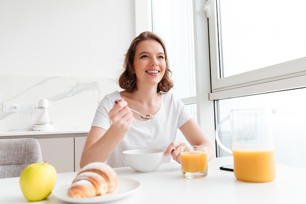 Portret van gelukkige vrouw die in witte t-shirt gezonde havermoutpap eten terwijl het zitten bij keukenlijst