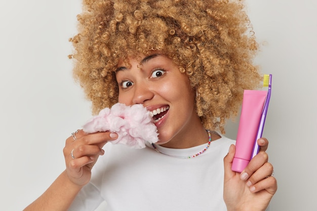 Portret van gelukkige vrouw met krullend haar eet suikerspin die schadelijk is voor je tanden houdt tube tandpasta en tandenborstel heeft blije expressie poses tegen witte achtergrond Mondhygiëne en zoet voedsel