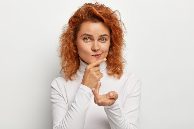 Gratis foto portret van gember vrouwtje strekt zich uit palm, portemonnees lippen en kin houdt close-up, ziet er recht uit, heeft natuurlijke schoonheid, gekleed in witte coltrui, beslist iets, vormt binnen