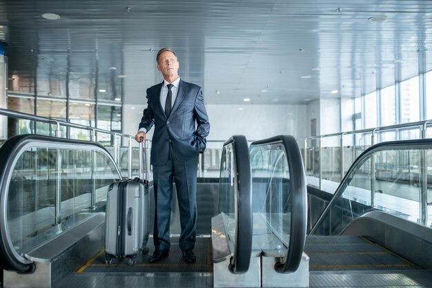 Portret van gemiddelde lengte van een serieuze, peinzende man met de trolleykoffer die van de roltrap stapt