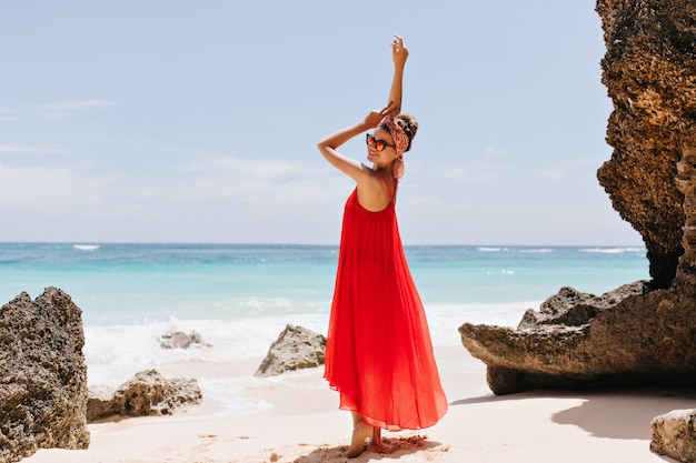 Portret van gemiddelde lengte van elegante Europese vrouw die dichtbij oceaan glimlacht. Prachtig gelooid meisje in het rood poseren op het strand