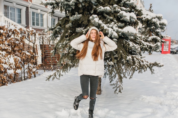 Portret van gemiddelde lengte van ontspannen blonde vrouw in zwarte broek dansen op besneeuwde straat met glimlach. buiten foto van grappige sierlijke vrouw poseren met handen omhoog voor groene sparren in winterdag.