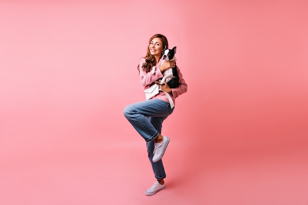 Portret van gemiddelde lengte van schattig meisje in trendy jeans met Franse bulldog. Dromerige blanke vrouw poseren met haar hond op roze.