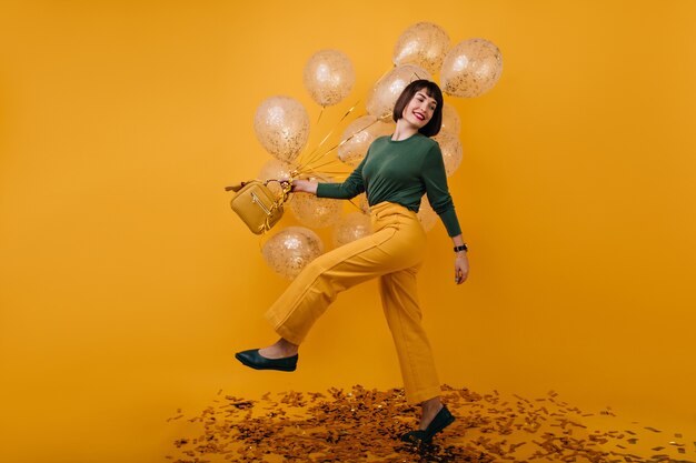 Portret van gemiddelde lengte van schattige vrouw dansen met feestballonnen. binnen schot van zalig donkerbruin meisje in gele broek met plezier op haar verjaardag.