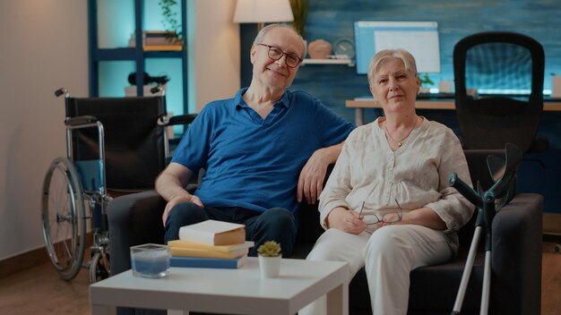 Portret van gepensioneerd echtpaar met een handicap zittend op de bank, kijkend naar de camera. Oudere man en vrouw naast krukken en rolstoel voor toegankelijkheid en mobiliteit, genietend van vrije tijd.