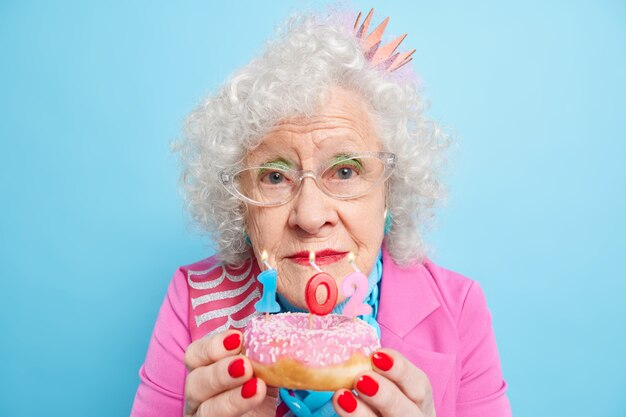 Portret van gerimpelde vrouw met krullend grijs haar houdt geglazuurde donut vast met nummerkaarsen viert 102e verjaardag heeft rode nagels draagt make-up looks direct,