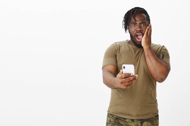 Portret van geschokte man in een bruin t-shirt poseren tegen de witte muur met zijn telefoon