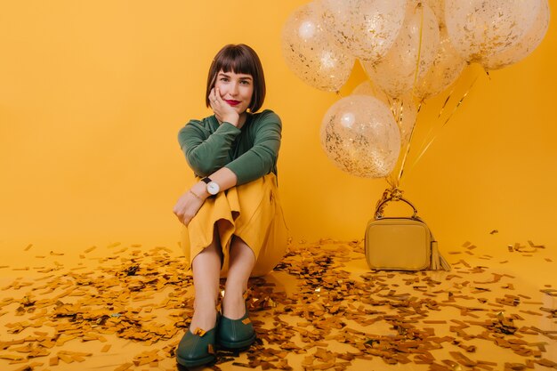 portret van geweldige jonge vrouw met kort haar, zittend op sparkle confetti. Indoor foto van enthousiast vrouwelijk model in groene trui poseren na feest.