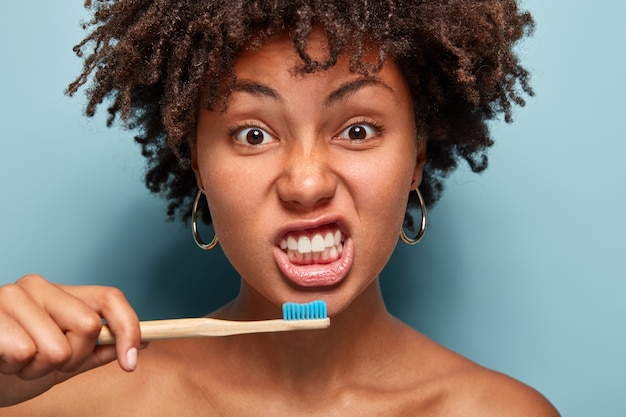 Portret van gezond meisje tanden poetsen, houten borstel houdt, heeft ochtendroutine, krullend haar, vormt binnen over blauwe muur, toont blote schouders vroeg wakker. Mensen, etniciteit en hygiëneconcept