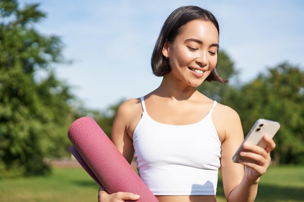 Portret van glimlachende aziatische vrouw die met yogamat naar haar smartphone kijkt en een applicatie leest die in het park staat en een sportuniform draagt