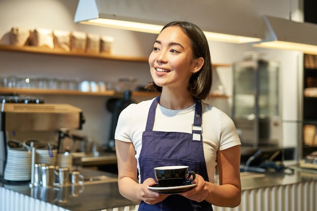 Portret van glimlachende aziatische vrouw in schortbarista die u kop van koffie geeft die in koffie werkt die dr