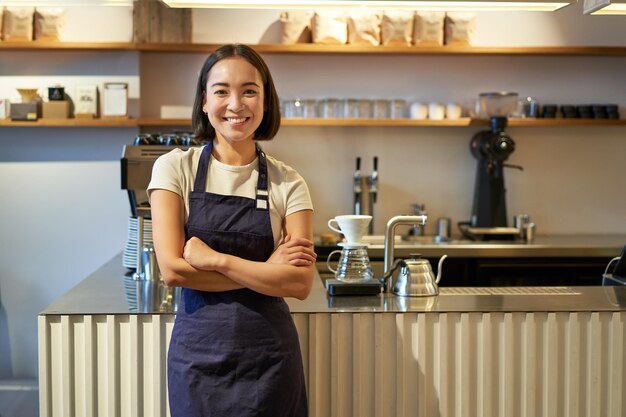 Portret van glimlachende aziatische vrouwelijke barista die schort draagt die zich dichtbij toonbank bevindt met koffie die binnen werkt