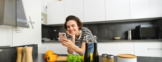 Portret van glimlachende jonge vrouw die in de keuken kookt en video's kijkt op een smartphone-app