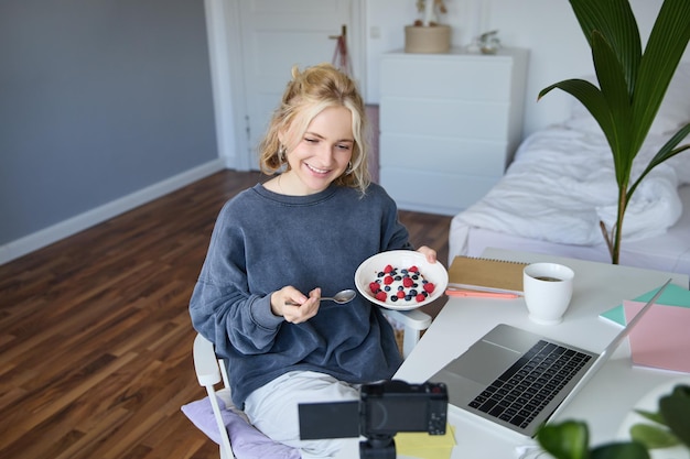Gratis foto portret van glimlachende jonge vrouwelijke vlogger die inhoud opneemt en creëert in haar kamer terwijl ze voorin eet