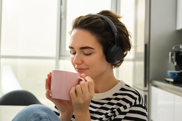 Portret van glimlachende ontspannen vrouw drinkt warme thee en luistert naar muziek of ebook in de keuken