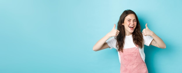 Portret van glimlachende vrolijke jonge vrouw met krullend kapsel die duimen omhoog laat zien in goedkeuring zeg ja geef aanbeveling lof goede zaak die tegen blauwe achtergrond staat