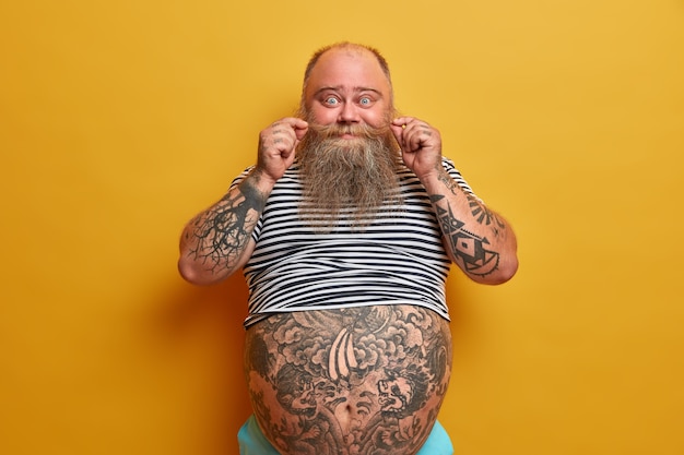 Gratis foto portret van grappige bebaarde man krult snor, heeft getatoeëerde armen en buik, gekleed in ondermaats gestreept mouwloos t-shirt, heeft probleem van obesitas en overgewicht, geïsoleerd op gele muur