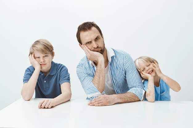 Portret van grappige familie van vader en zonen die aan tafel zitten, hoofd aan kant leunend en gezichten trekken