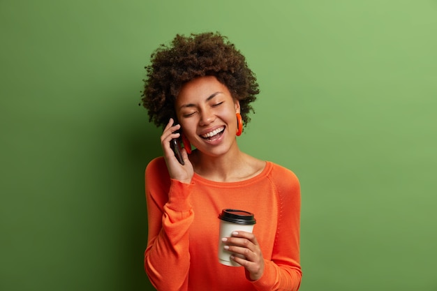 Portret van grappige vrouw met krullend haar, lacht gelukkig, heeft telefoongesprek, wordt geamuseerd door vriend, drinkt koffie uit wegwerpbeker, terloops gekleed, sluit de ogen