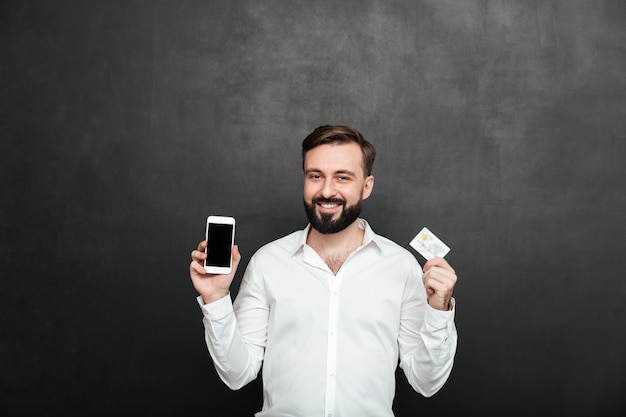 Portret van het donkerbruine mens stellen op camera die smartphone en creditcard voor online winkelen gebruiken, geïsoleerd over donkergrijs