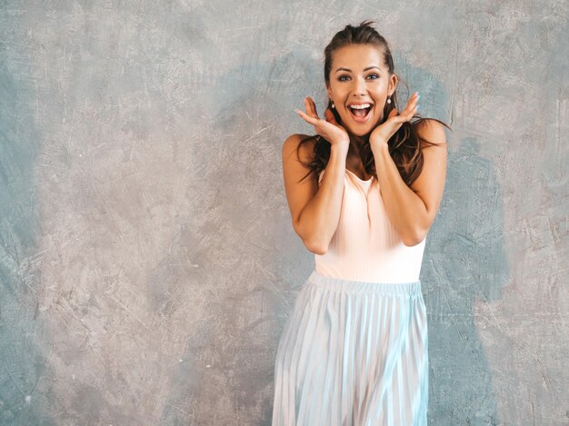 Portret van het jonge mooie verraste vrouw kijken met handen dichtbij gezicht. Trendy meisje in casual zomerkleding. Geschokt vrouwelijke poseren in de buurt van grijze muur