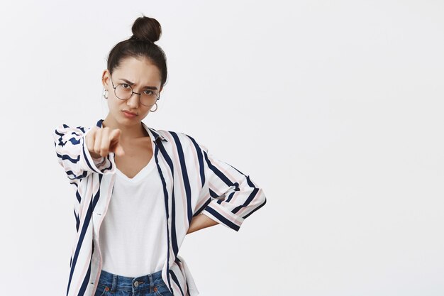 Portret van humeurig ontevreden aantrekkelijk vrouwelijk model in overhemd en glazen, wijzend, hand houdend op heup en fronsen van teleurstelling