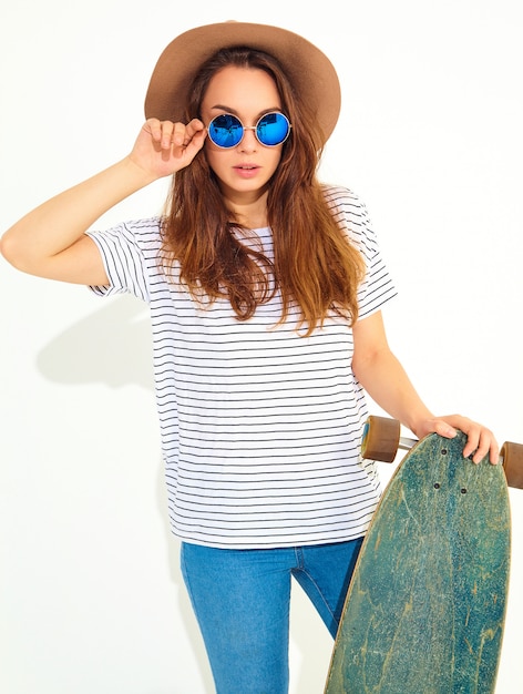 Portret van jong modieus vrouwenmodel in toevallige de zomerkleren in het bruine hoed stellen met longboardbureau. geïsoleerd op wit