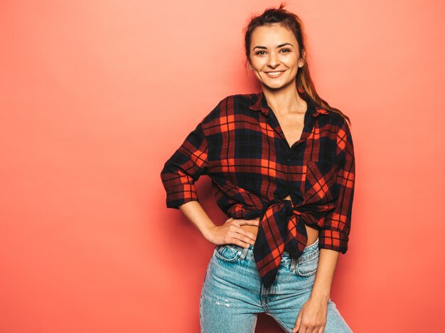 Portret van jong mooi glimlachend hipster meisje in trendy de zomer geruit overhemd en jeanskleren. Het sexy onbezorgde vrouw stellen dichtbij roze muur in studio. Positief model zonder make-up