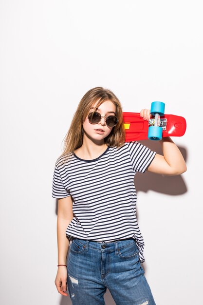Portret van jong tienermeisje in zonnebril die met skateboard stellen terwijl status over witte muur