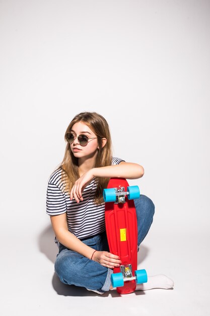 Portret van jong tienermeisje in zonnebril die met skateboard stellen terwijl status over witte muur