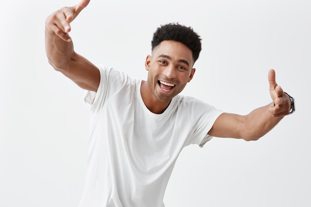 Portret van jonge aantrekkelijke donkere man met afro kapsel in witte casual t-shirt lachend met tanden, gebarend met de handen in de camera, in de camera kijken met blij en opgewonden uitdrukking