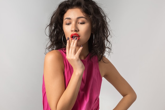 Portret van jonge aantrekkelijke exotische gelooide vrouw in roze stijlvolle jurk, rode lippenstift, sexy, verleidelijk, geïsoleerd, zomerseizoen stijl, modetrend, hand houdend op haar lippen, krullend haar