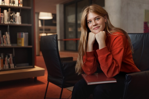 Portret van jonge aantrekkelijke roodharige vrouwelijke student, zit op een leunstoel die op wapens leunt en glimlacht.