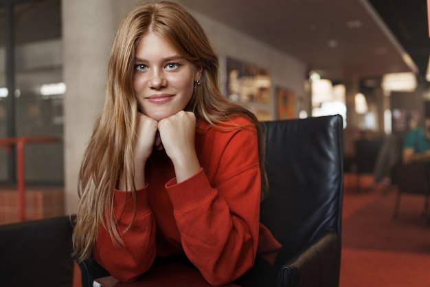 Portret van jonge aantrekkelijke roodharige vrouwelijke student, zit op een leunstoel die op wapens leunt en glimlacht.