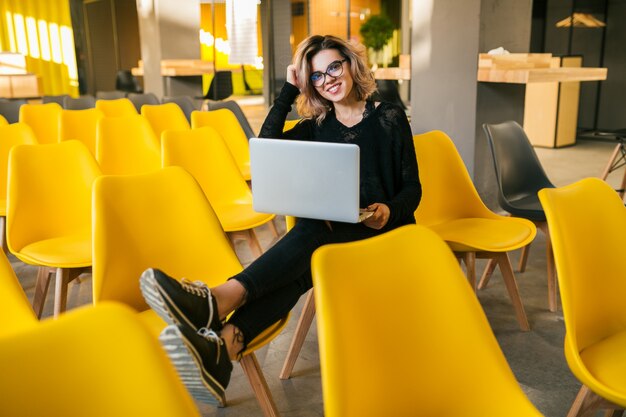 Portret van jonge aantrekkelijke vrouw zitten in de collegezaal, die op laptop werkt, bril, klaslokaal, veel gele stoelen, student leren, onderwijs online